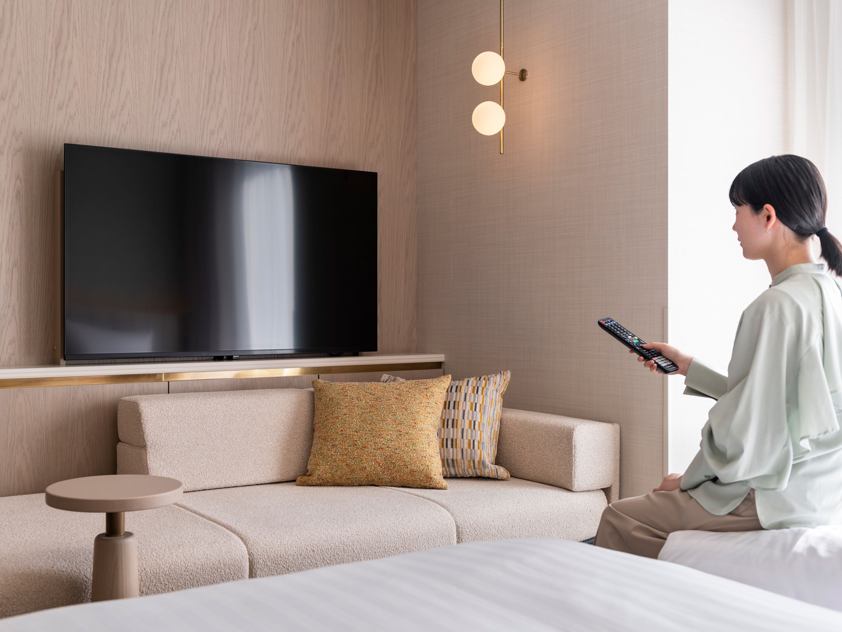 A modern hotel room with a large flat-screen TV mounted on the wall, a sofa with patterned cushions, a small round side table, and a remote control in hand.