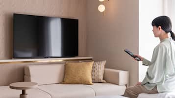 A modern hotel room with a large flat-screen TV mounted on the wall, a sofa with patterned cushions, a small round side table, and a remote control in hand.