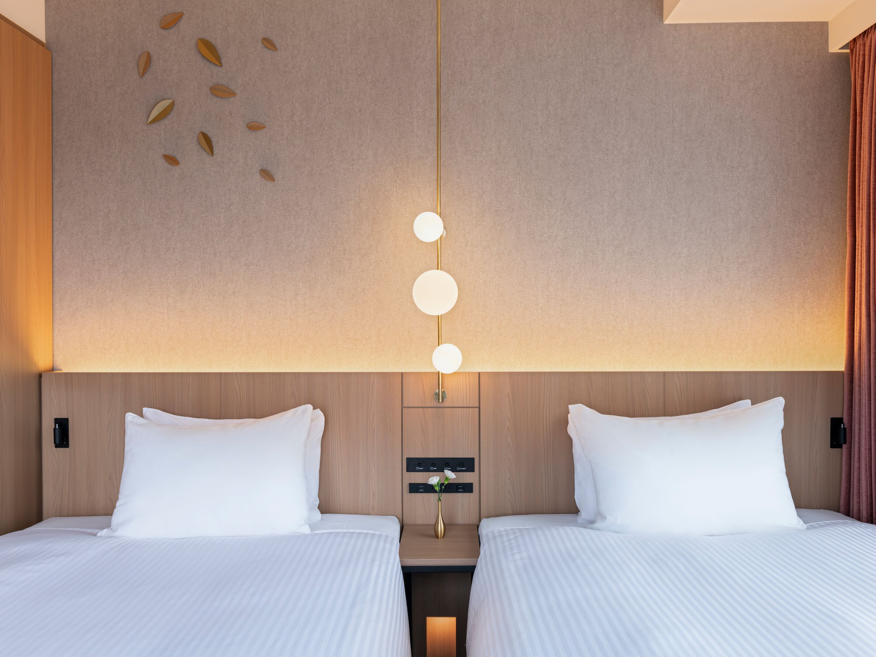 A hotel room with two beds, a wall-mounted light fixture, and a small table with a vase.
