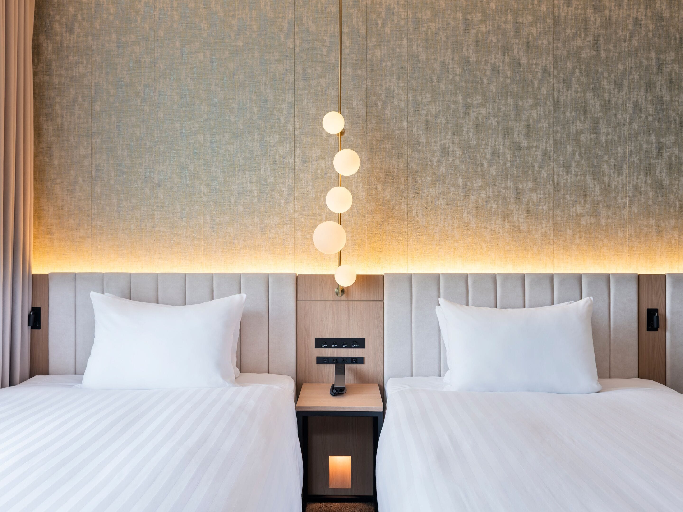 A hotel room with two beds, a nightstand with a lamp, and a wall-mounted light fixture.