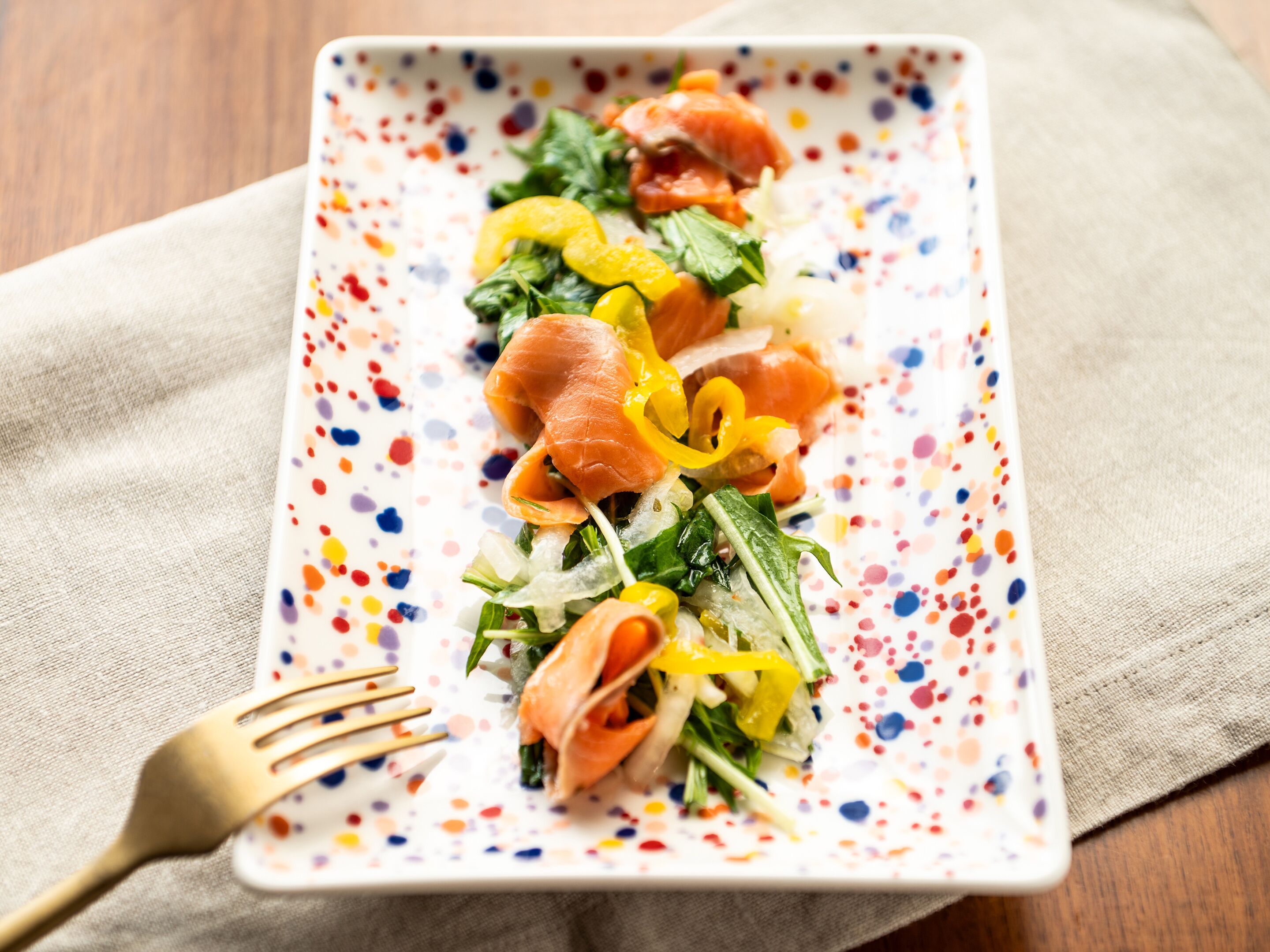 Prato retangular com um padrão colorido e salpicado, contendo uma salada de folhas verdes, pimentões amarelos e fatias de salmão, acompanhado por um garfo dourado sobre um guardanapo bege.