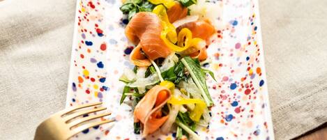 Prato retangular com um padrão colorido e salpicado, contendo uma salada de folhas verdes, pimentões amarelos e fatias de salmão, acompanhado por um garfo dourado sobre um guardanapo bege.