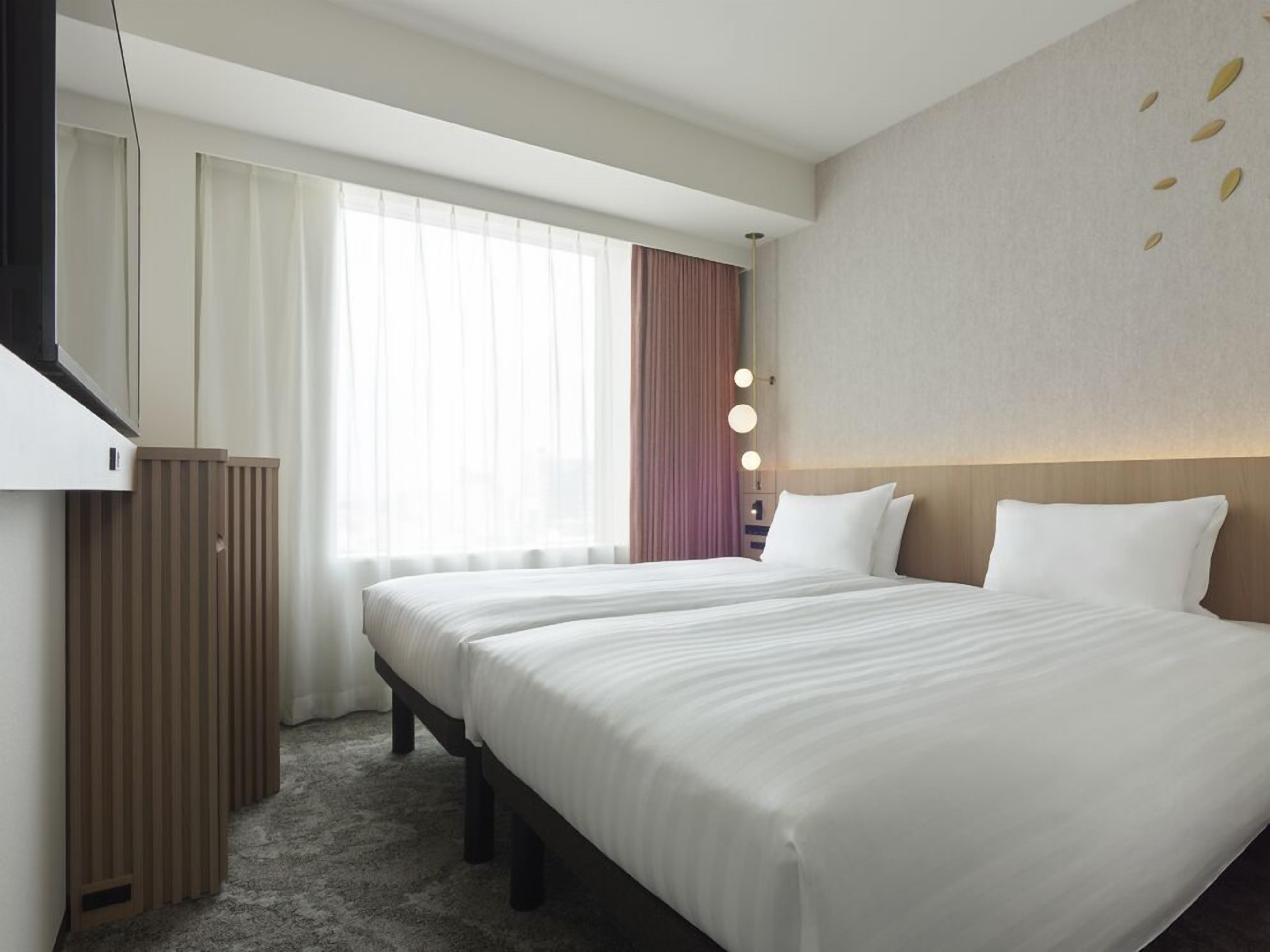 Habitación de hotel con dos camas, un ventanal con cortinas translúcidas, una mesita de noche con lámpara y un aplique de pared.
