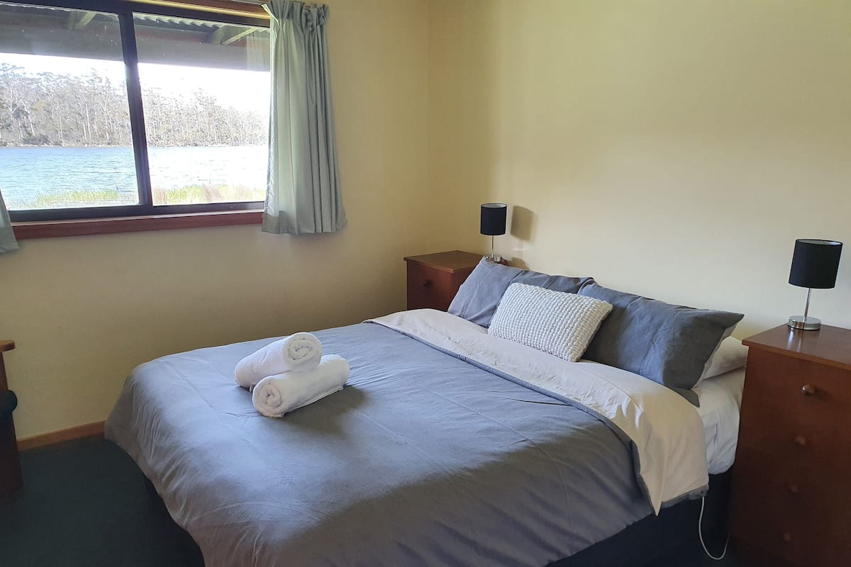 Una cama bien hecha con colcha azul y blanca, dos almohadas y dos toallas enrolladas. Se ve una ventana con vistas a árboles y una masa de agua.