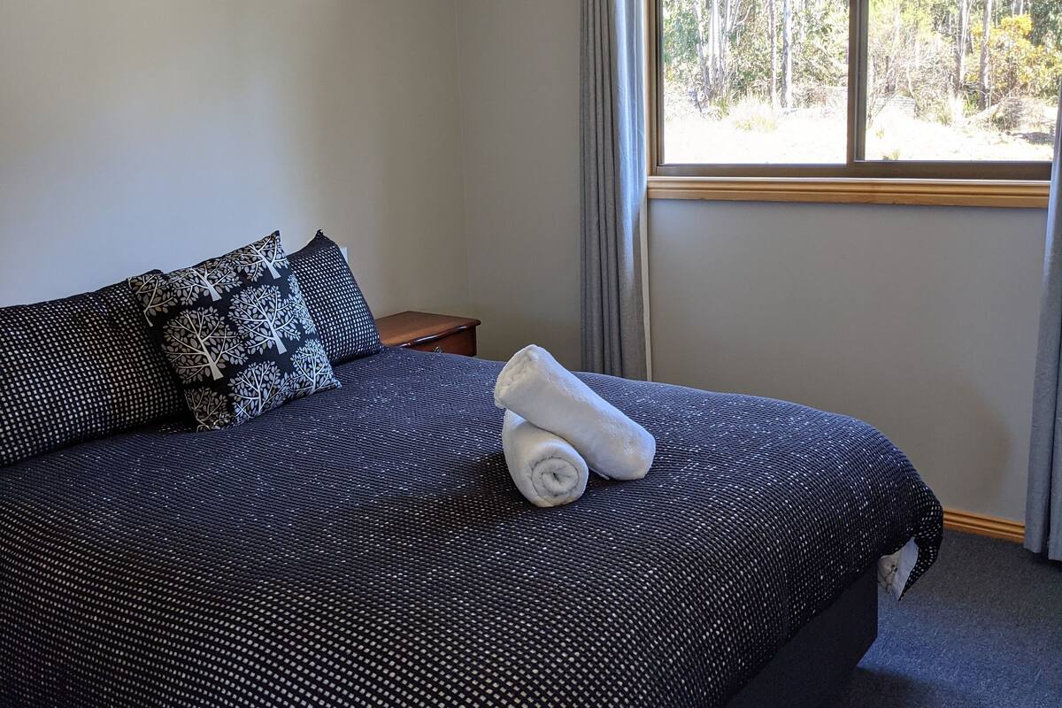 Una cama bien hecha con una colcha oscura estampada y dos almohadones decorativos, una ventana con vista a árboles y una mesita de noche con una lámpara.