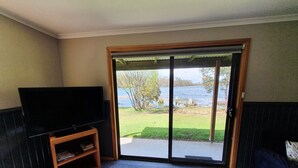 A living room with a flat-screen TV on a wooden stand, a sliding glass door leading to a grassy area with a lake view. - Lake Yalleena Nature Retreat (Lake Leake)