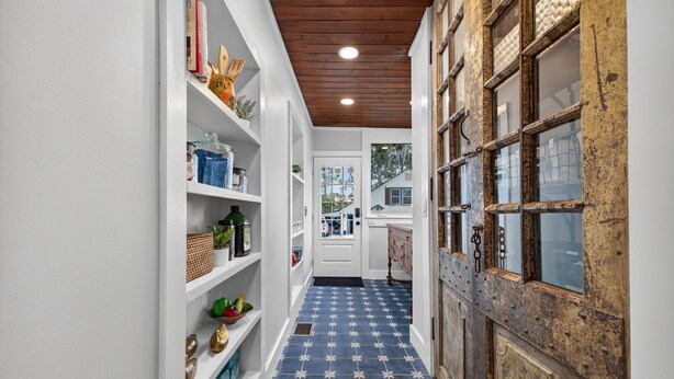 A narrow hallway with white walls, a blue patterned floor, and a rustic wooden door. Shelves on the left hold various items, and a white door at the end leads to another room. - The Estate at Mount Dora (Mount Dora)