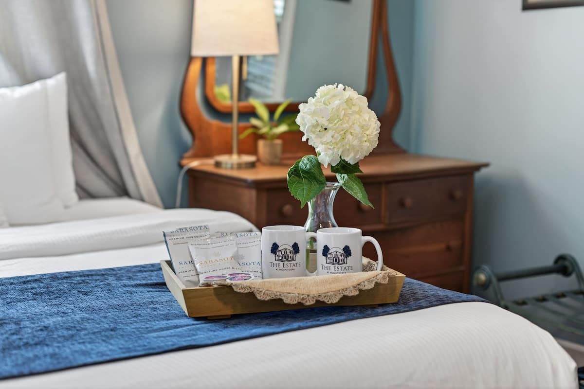 Ein ordentlich bezogenes Bett mit blauer Decke, ein Nachttisch aus Holz mit Lampe, eine Vase mit weißen Blumen und zwei Tassen mit Hotel-Logo.