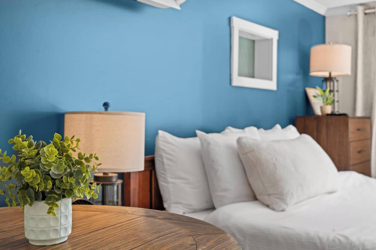 Ein Zimmer mit einem Bett, einem runden Holztisch mit einem Blumentopf, einer Lampe und einer Kommode.