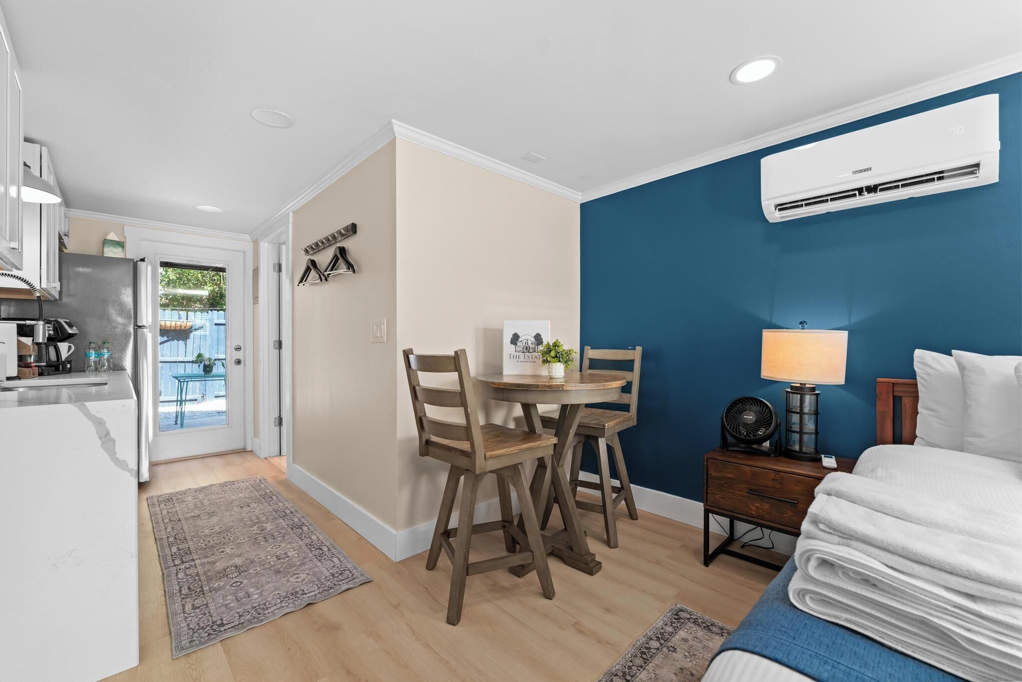 A compact room with a bed, a small table with chairs, a kitchen area, and a blue accent wall.