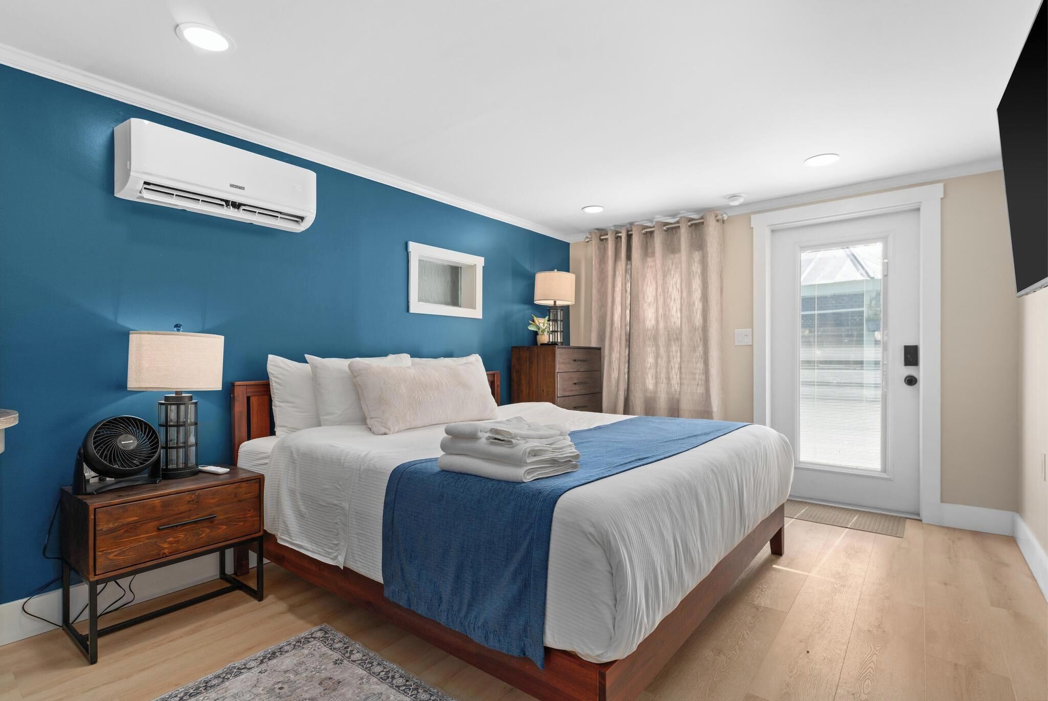 A bedroom with a bed, bedside tables, an air conditioner, a lamp, a mirror, and a view of the outdoors.