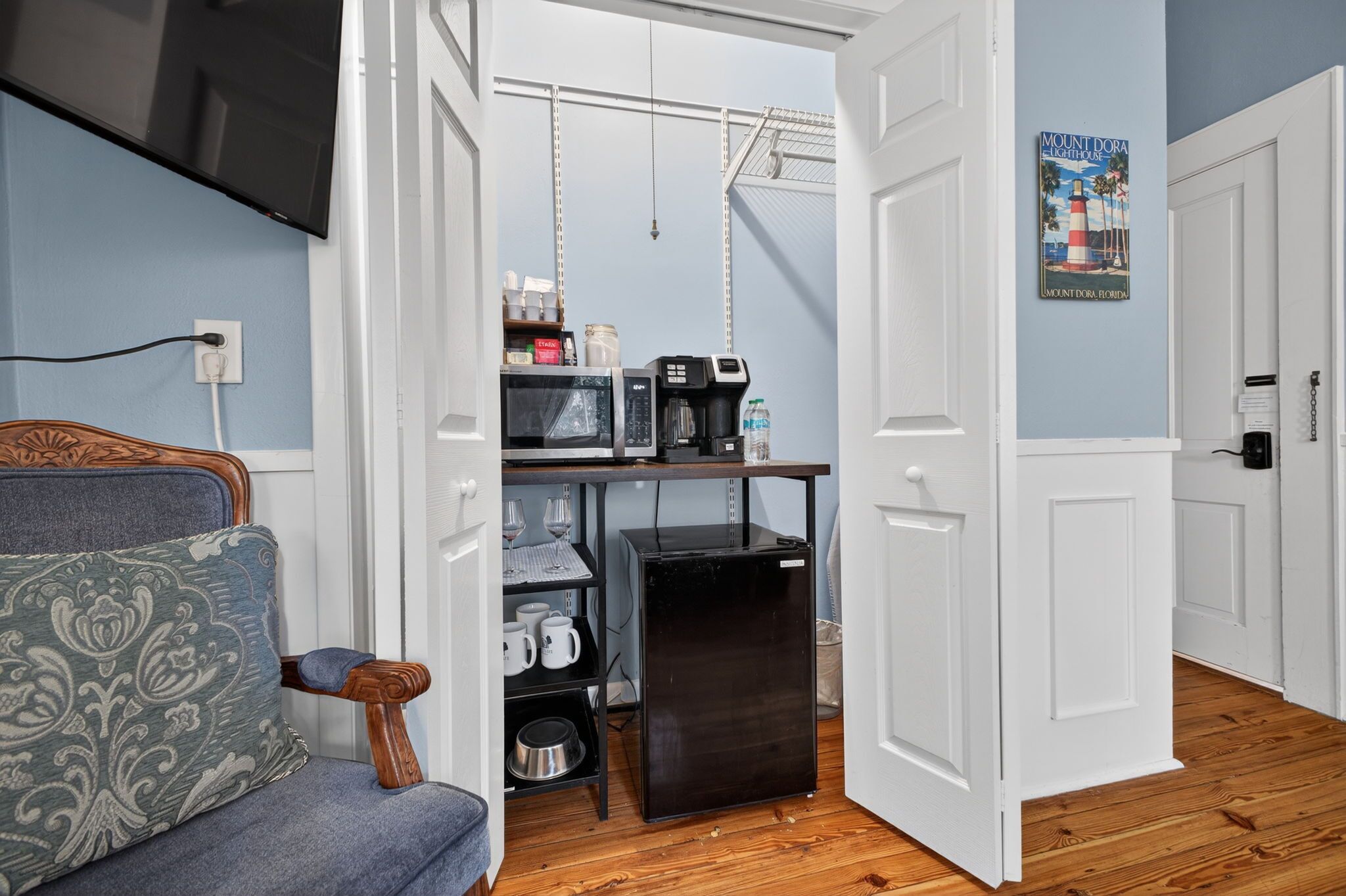 A compact room with a kitchenette, a television, a chair with a patterned cushion, and a poster on the wall.