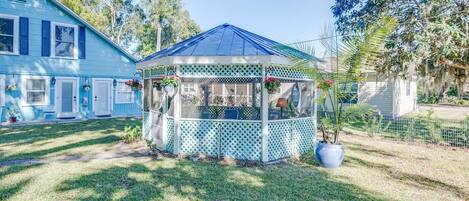 Un kiosque orné d’un treillis, entouré d’une pelouse bien entretenue et d’une maison aux volets bleus.