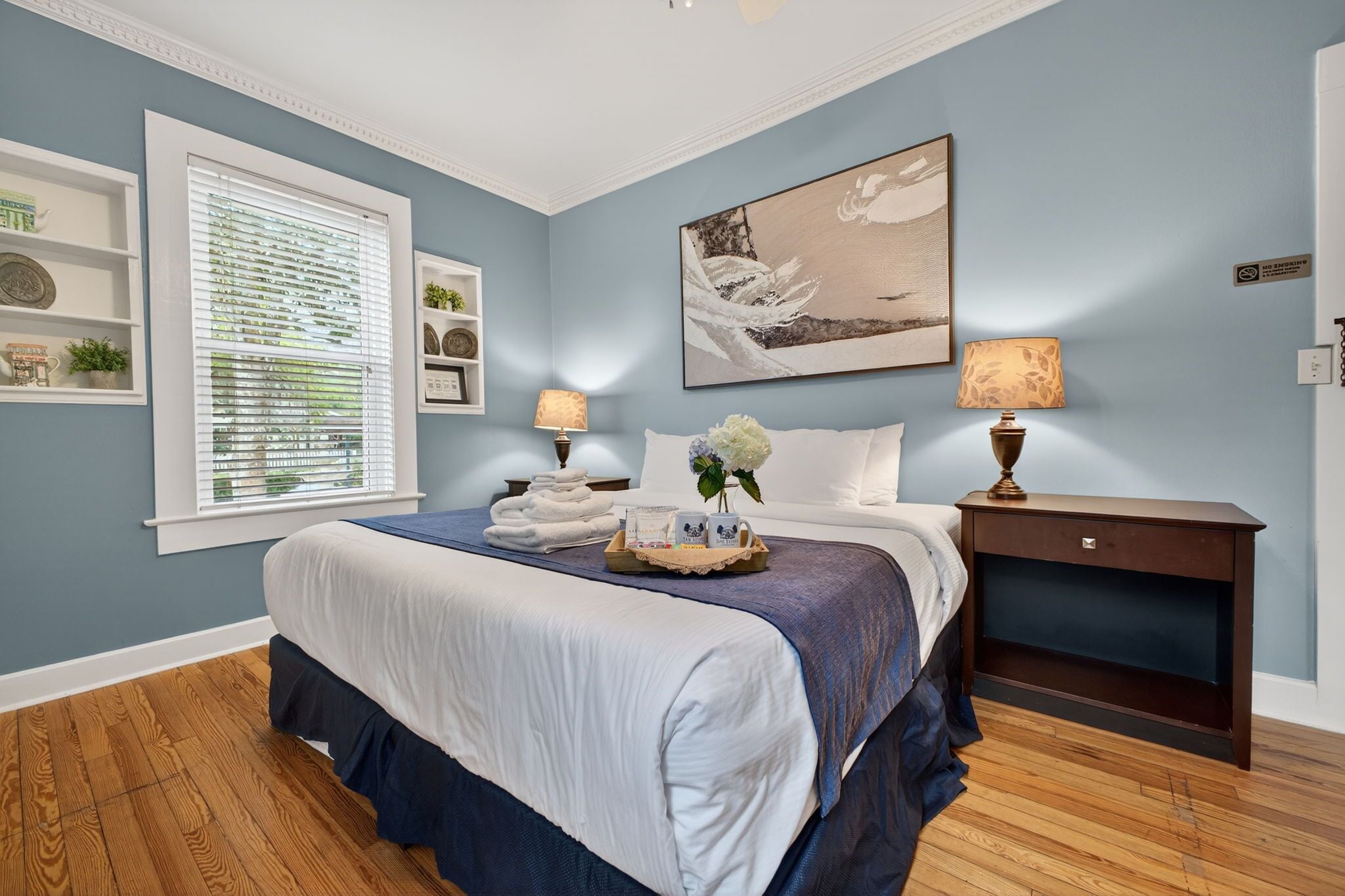 A bedroom with a bed, a nightstand, a lamp, a window with blinds, and a wall with a painting.