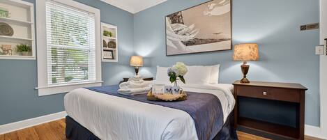 A bedroom with a bed, a nightstand, a lamp, a window with blinds, and a wall with a painting.
