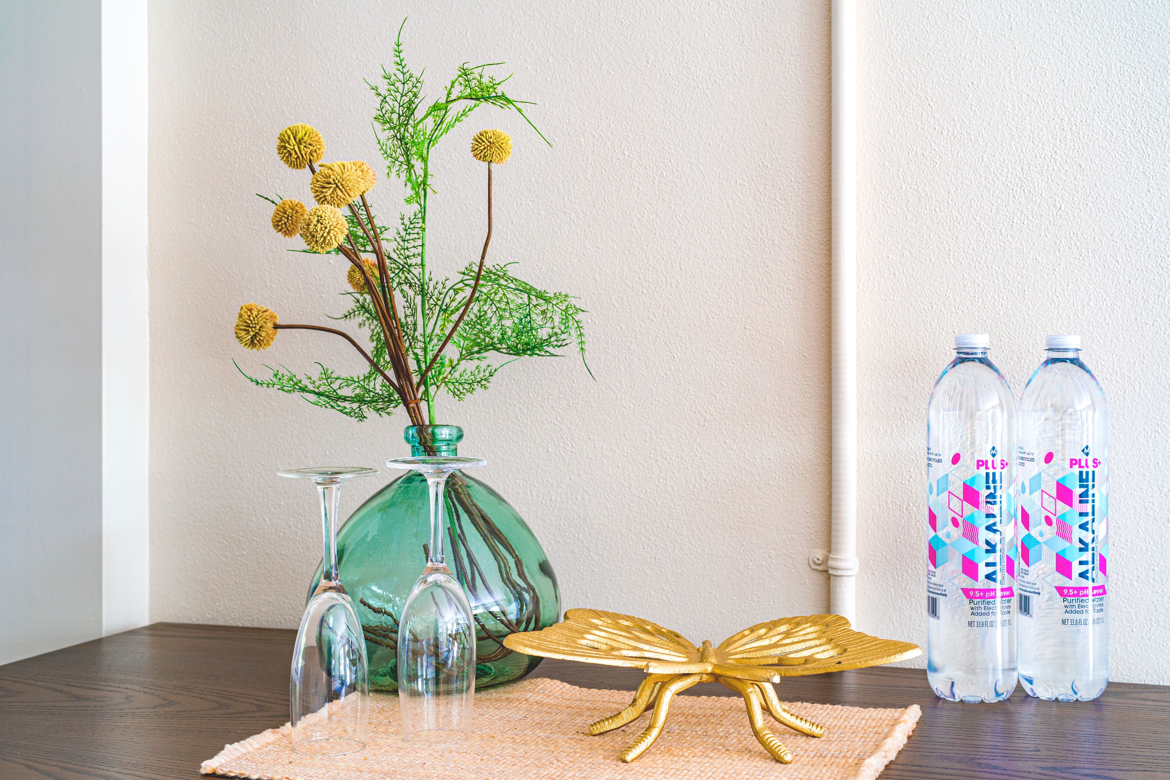 Una mesa con un jarrón verde con flores amarillas, dos copas de vino transparentes, una decoración de mariposa dorada y dos botellas de agua.