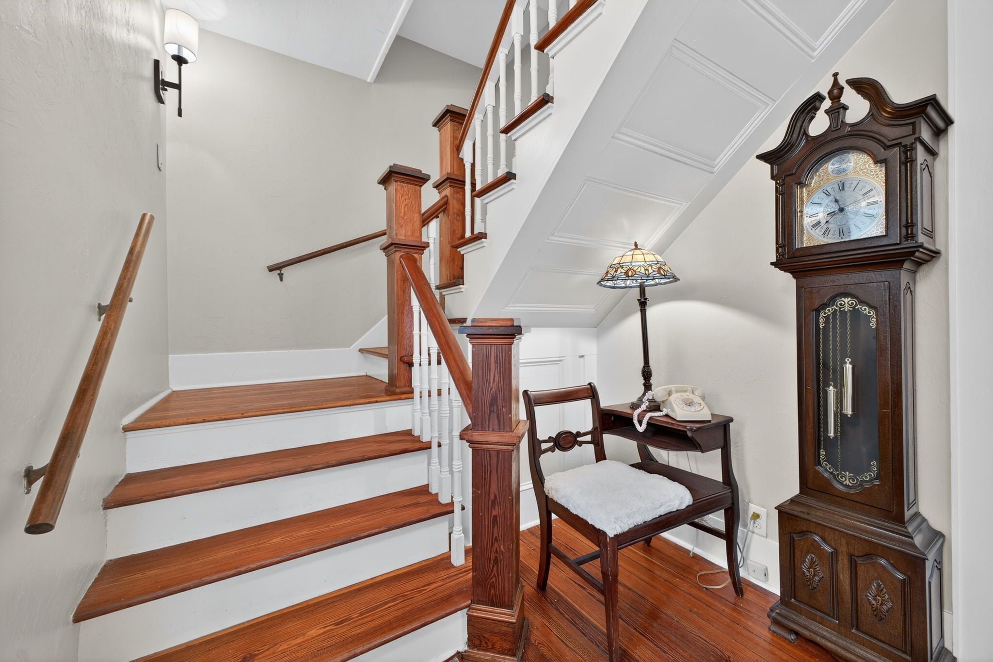 A staircase with wooden steps and white railings, a wooden chair, a telephone, and a tall grandfather clock.
