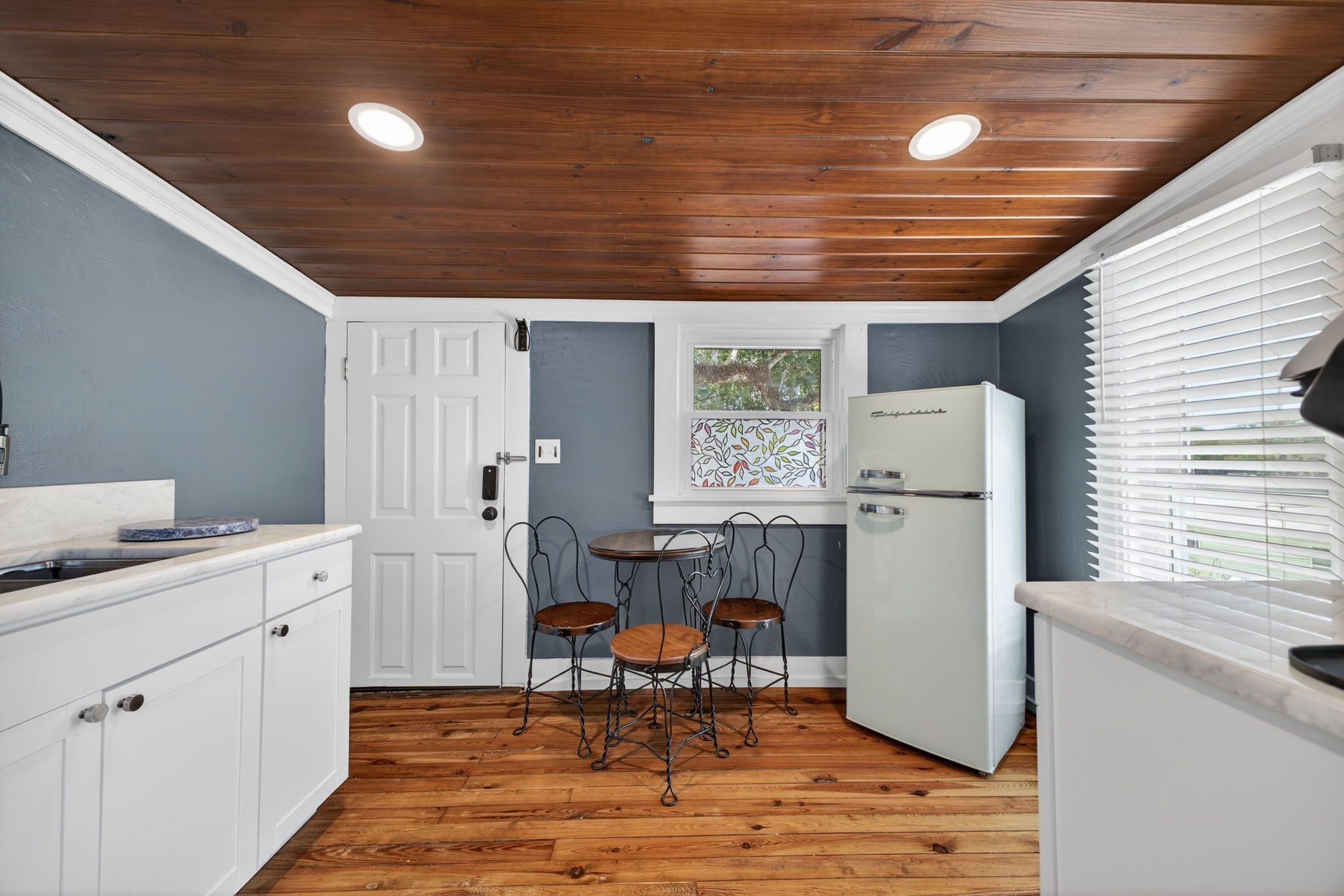 A compact kitchen with wooden flooring, white cabinetry, a small dining table with chairs, a refrigerator, and a window with blinds.