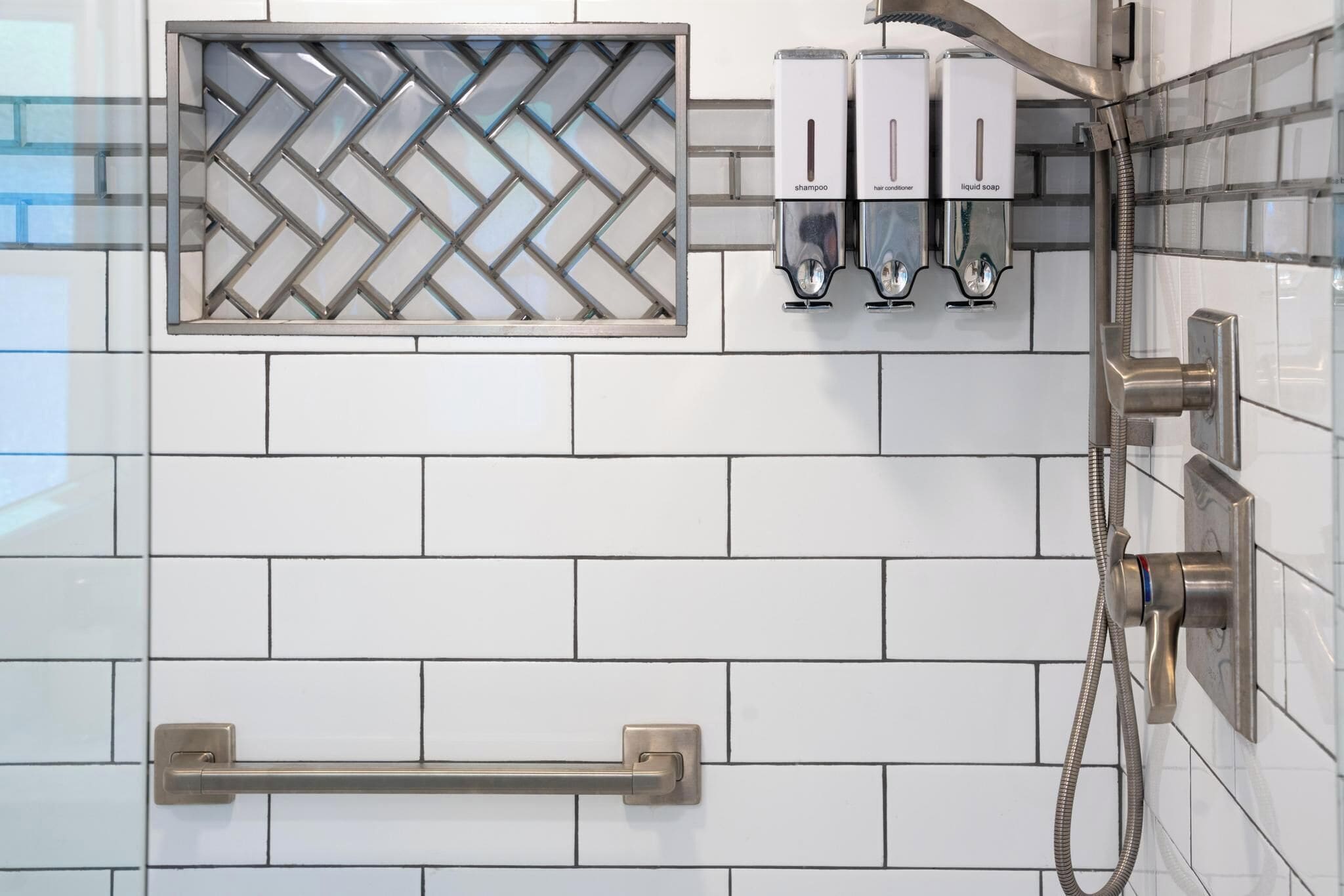 Un bagno con pareti piastrellate di bianco, una doccia con soffione manuale e tre dispenser per shampoo, balsamo e sapone liquido.