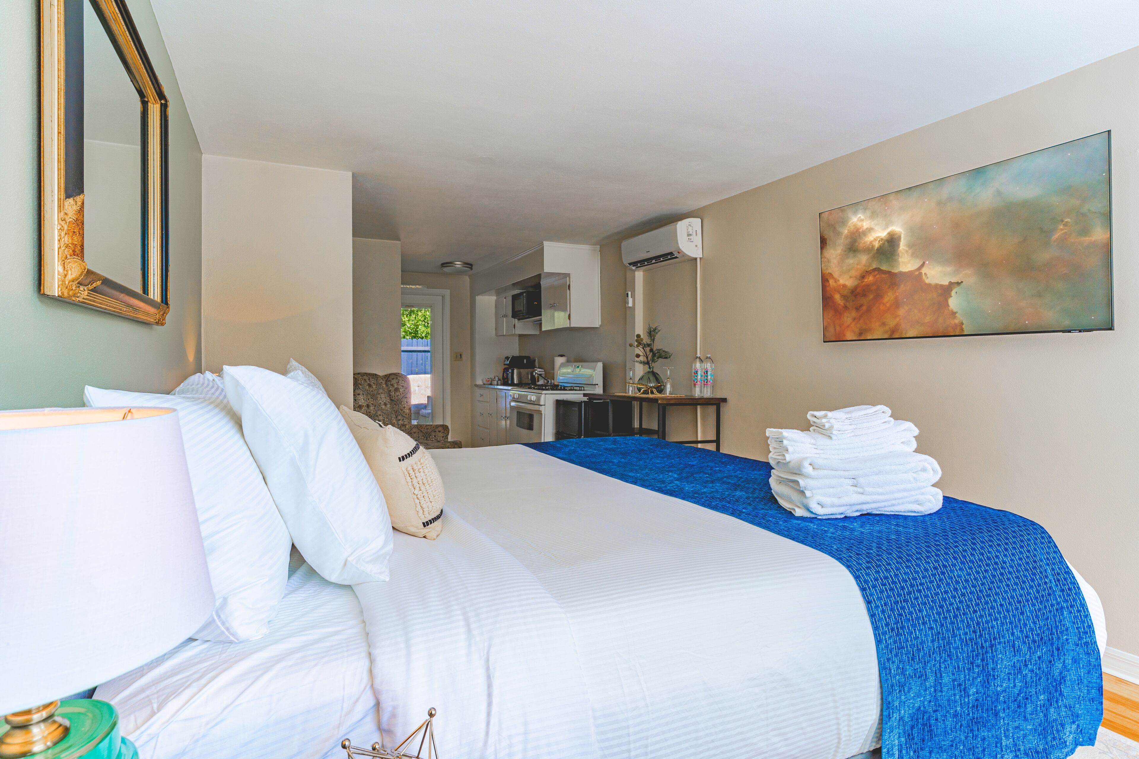 Een hotelkamer met een groot bed, een bureau met een computer, een kitchenette en een schilderij aan de muur.