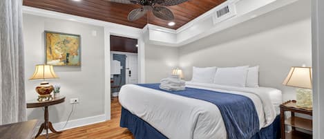 A bedroom with a wooden ceiling, a bed with white and blue bedding, a standing lamp, and a painting on the wall.
