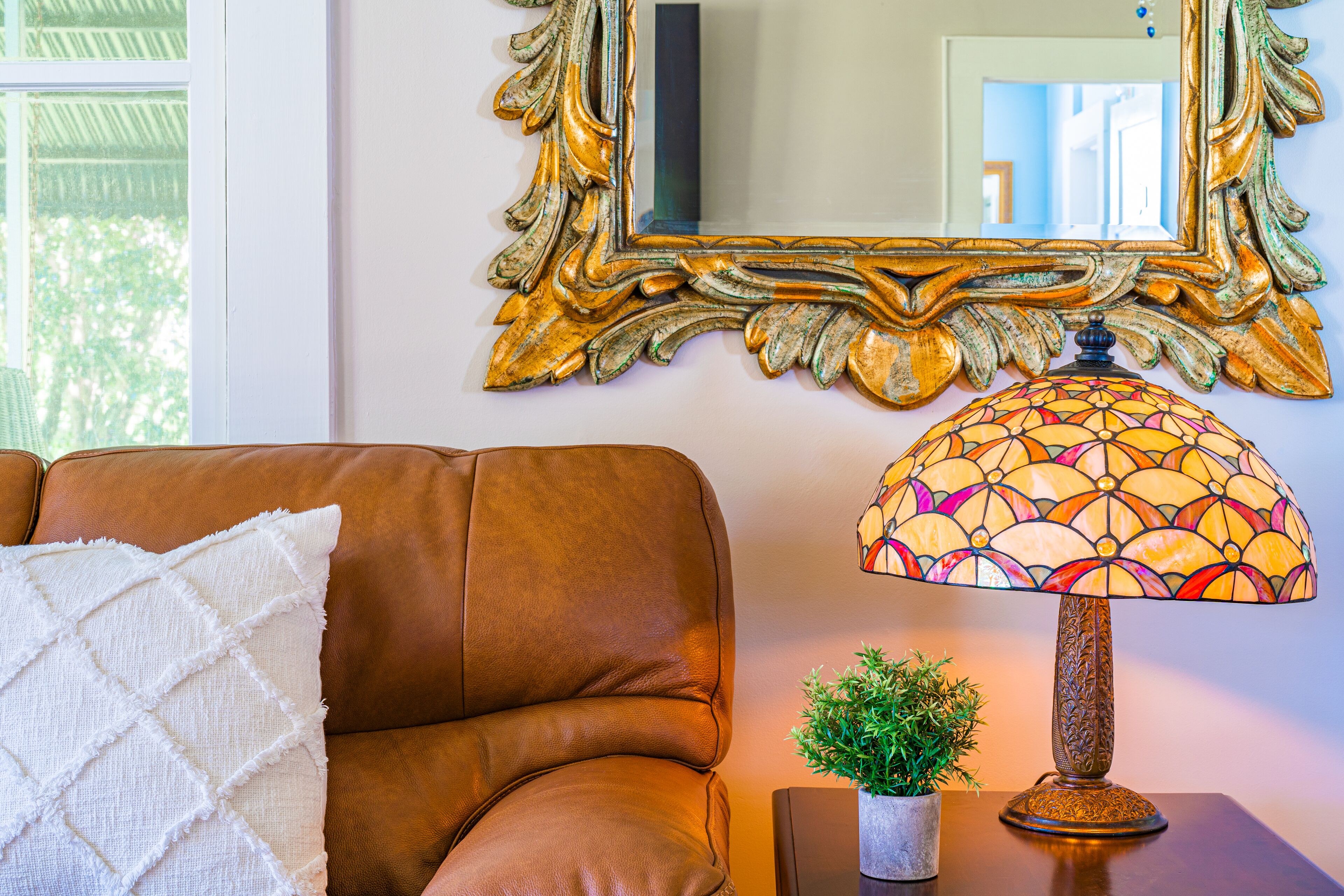 A brown leather sofa with a white quilted pillow, a stained glass lamp, and a small potted plant on a wooden table.