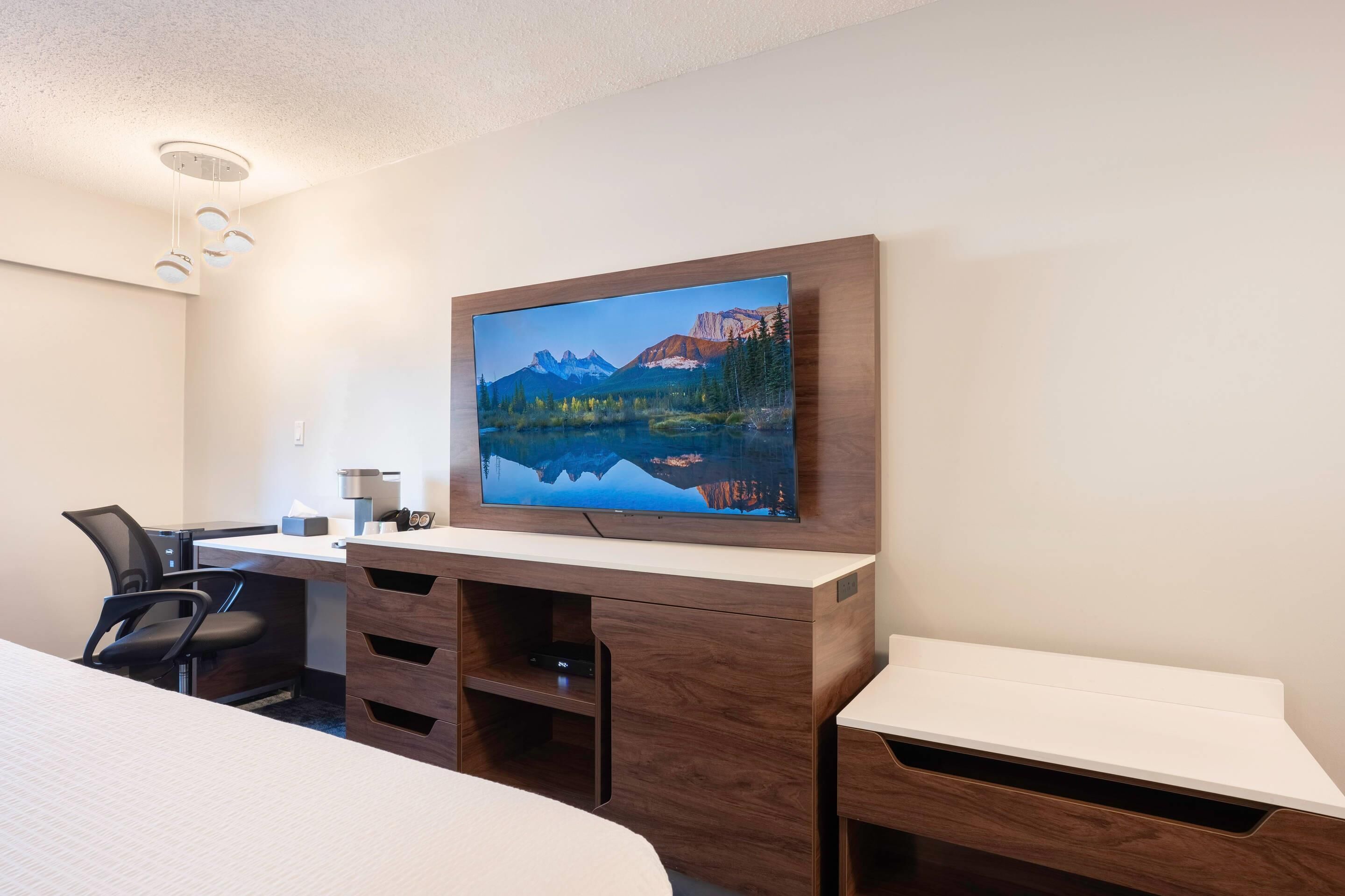 A hotel room with a bed, a desk with a computer, a TV mounted on the wall, and a view of mountains and a lake.