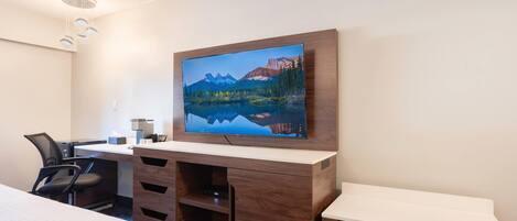 A hotel room with a bed, a desk with a computer, a TV mounted on the wall, and a view of mountains and a lake.