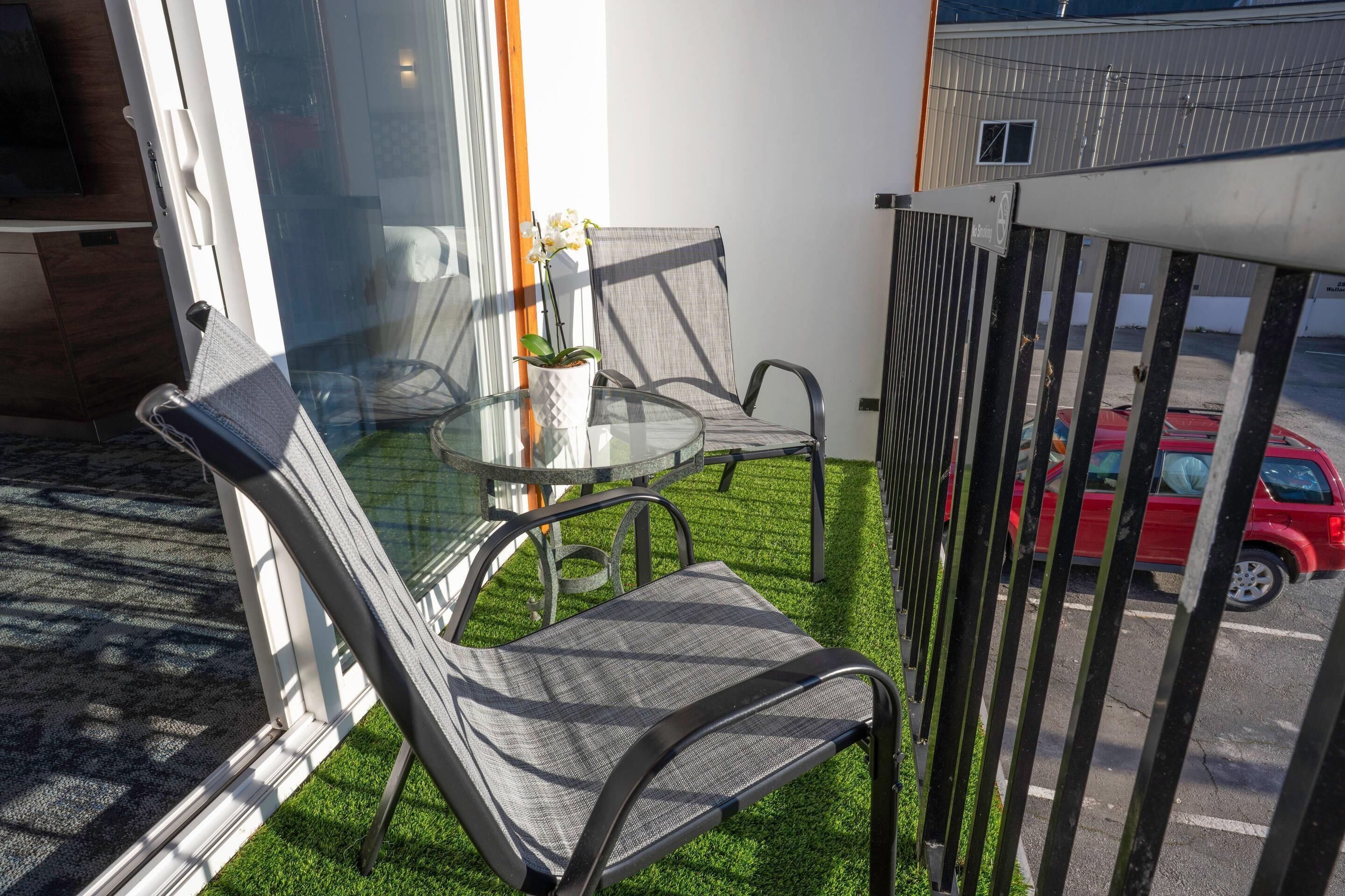 Un balcón con una mesa de vidrio, dos sillas, una planta en maceta y vista a un estacionamiento.