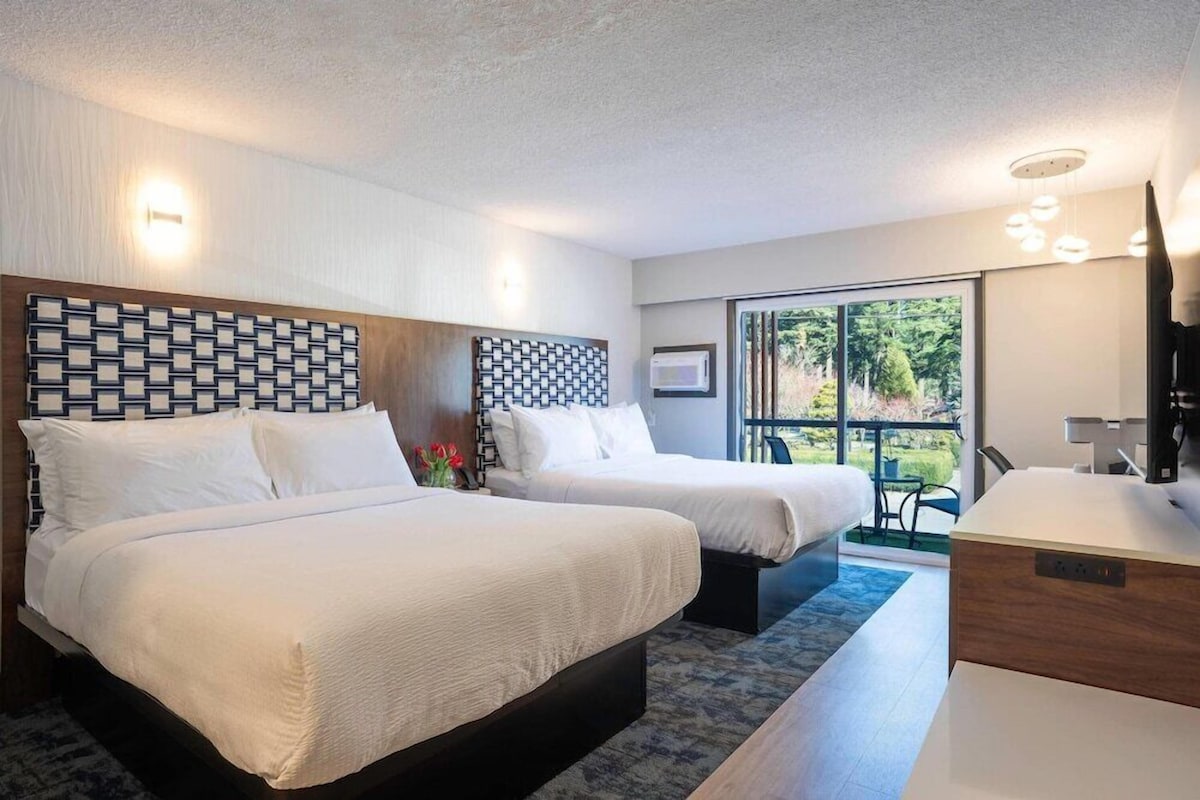 A hotel room with two beds, a desk, a chair, a large window, and a view of greenery outside.