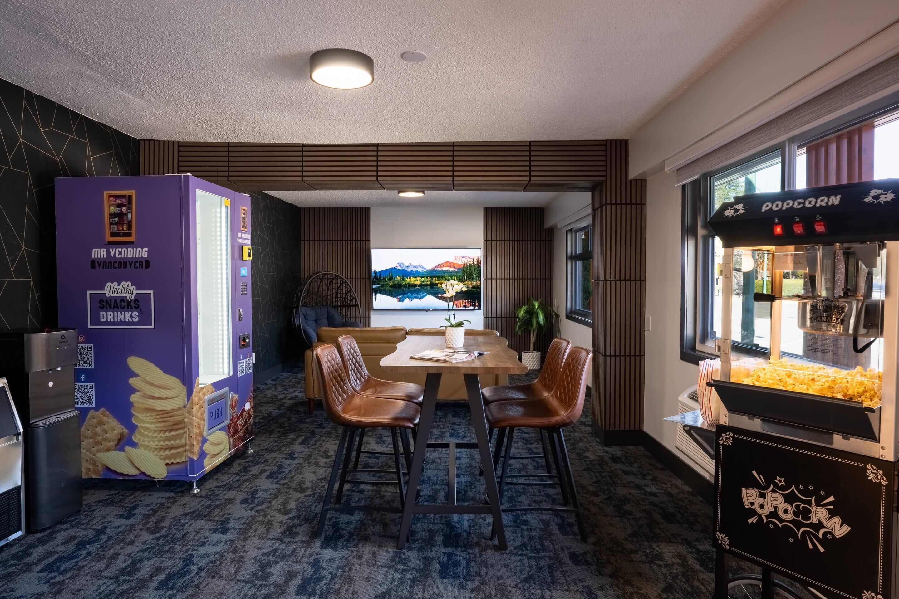 A modern dining area with a table and chairs, a vending machine, a popcorn machine, and a view of mountains through a window.