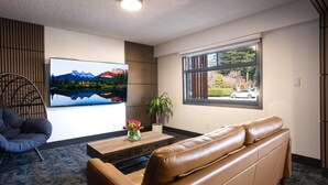 A modern living room with a leather sofa, a wooden coffee table, a large flat-screen TV, a potted plant, and a view of a mountain landscape through a window.