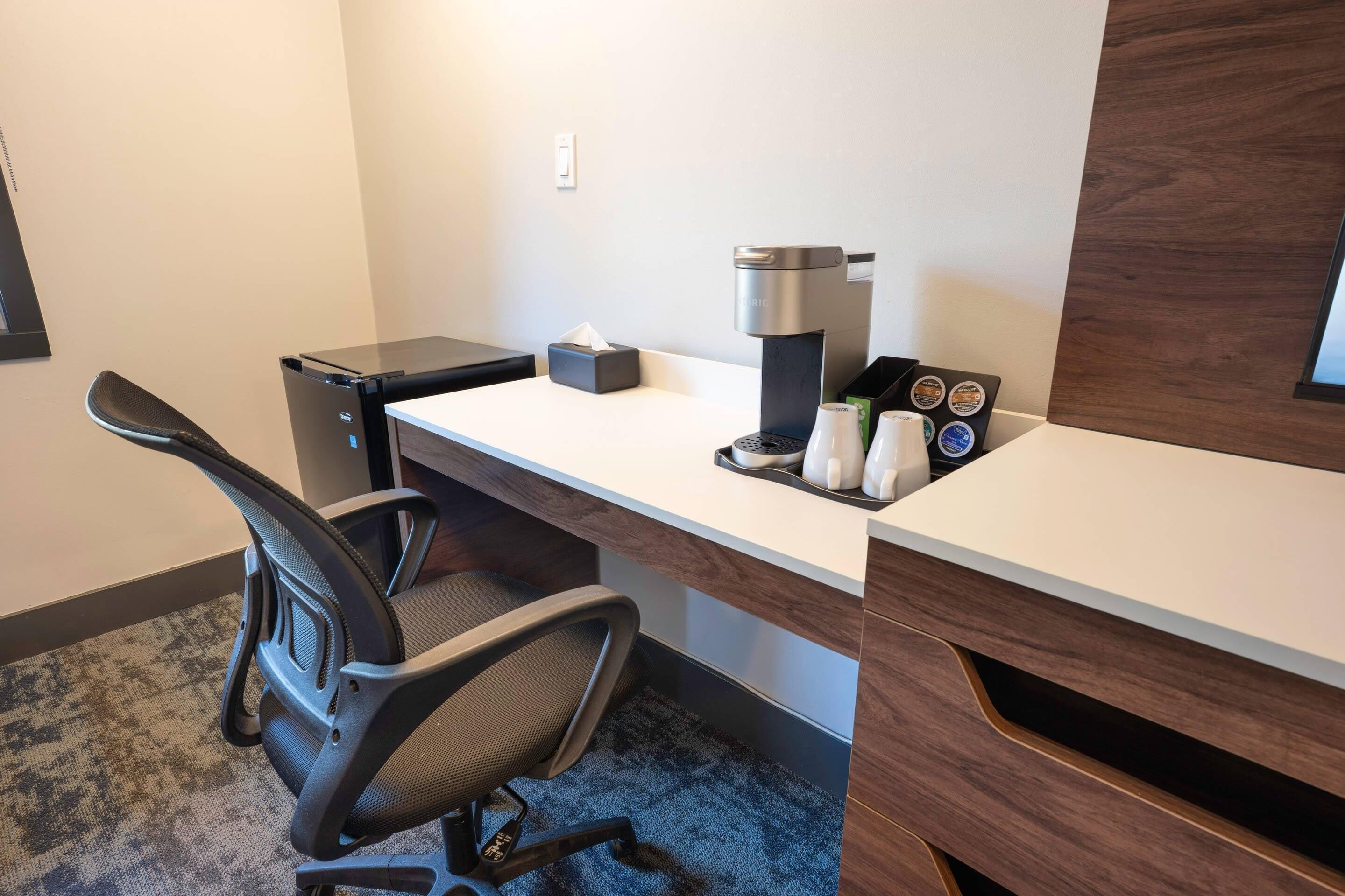 A hotel workspace with a desk, chair, coffee machine, tissue box, and a coffee pod holder.