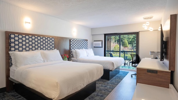 A hotel room with two beds, a desk, a chair, a TV, and a large window with a view of greenery.