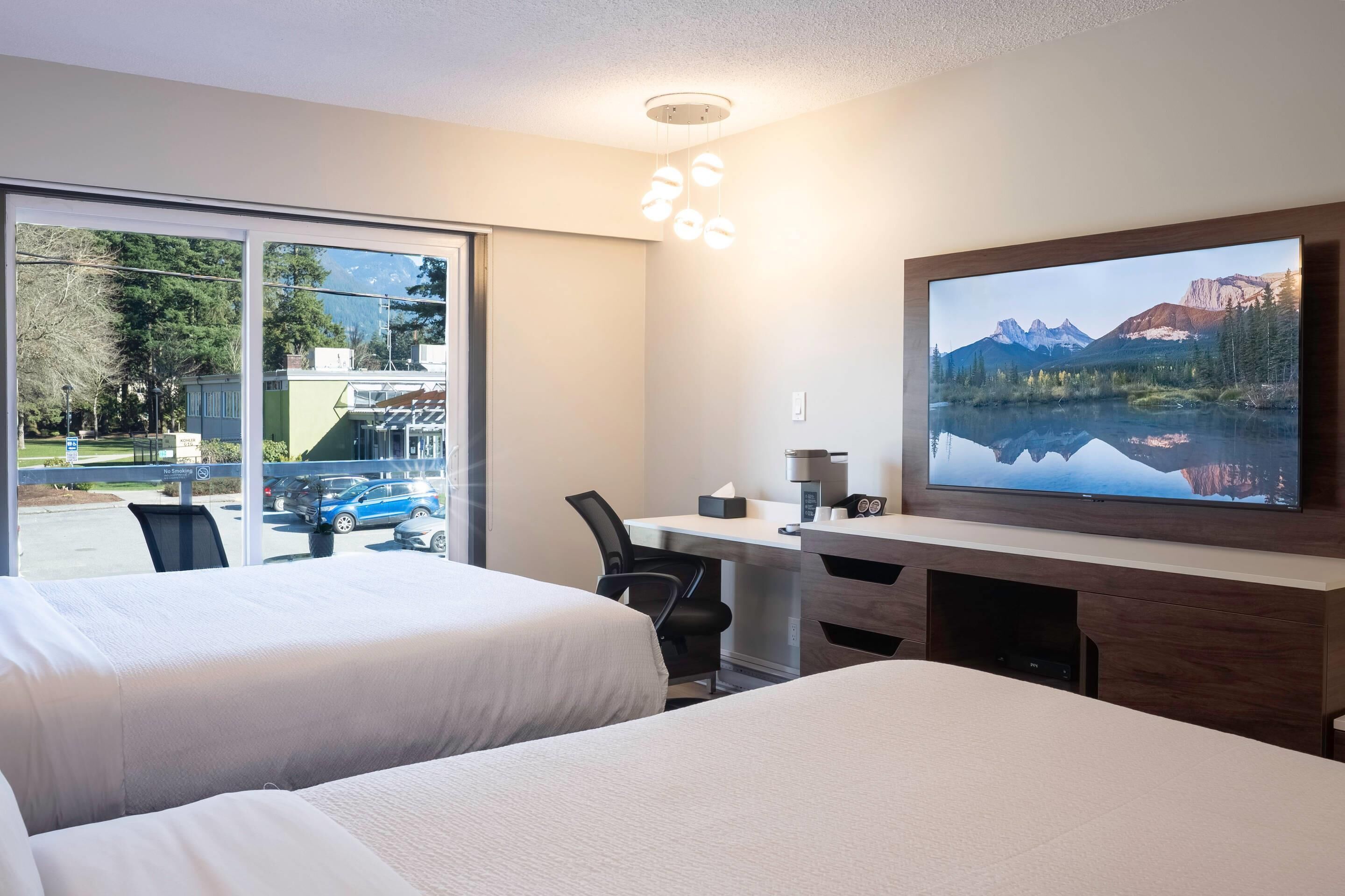 A hotel room with two beds, a desk, a chair, a TV, and a large window with a view of a parking lot and buildings.