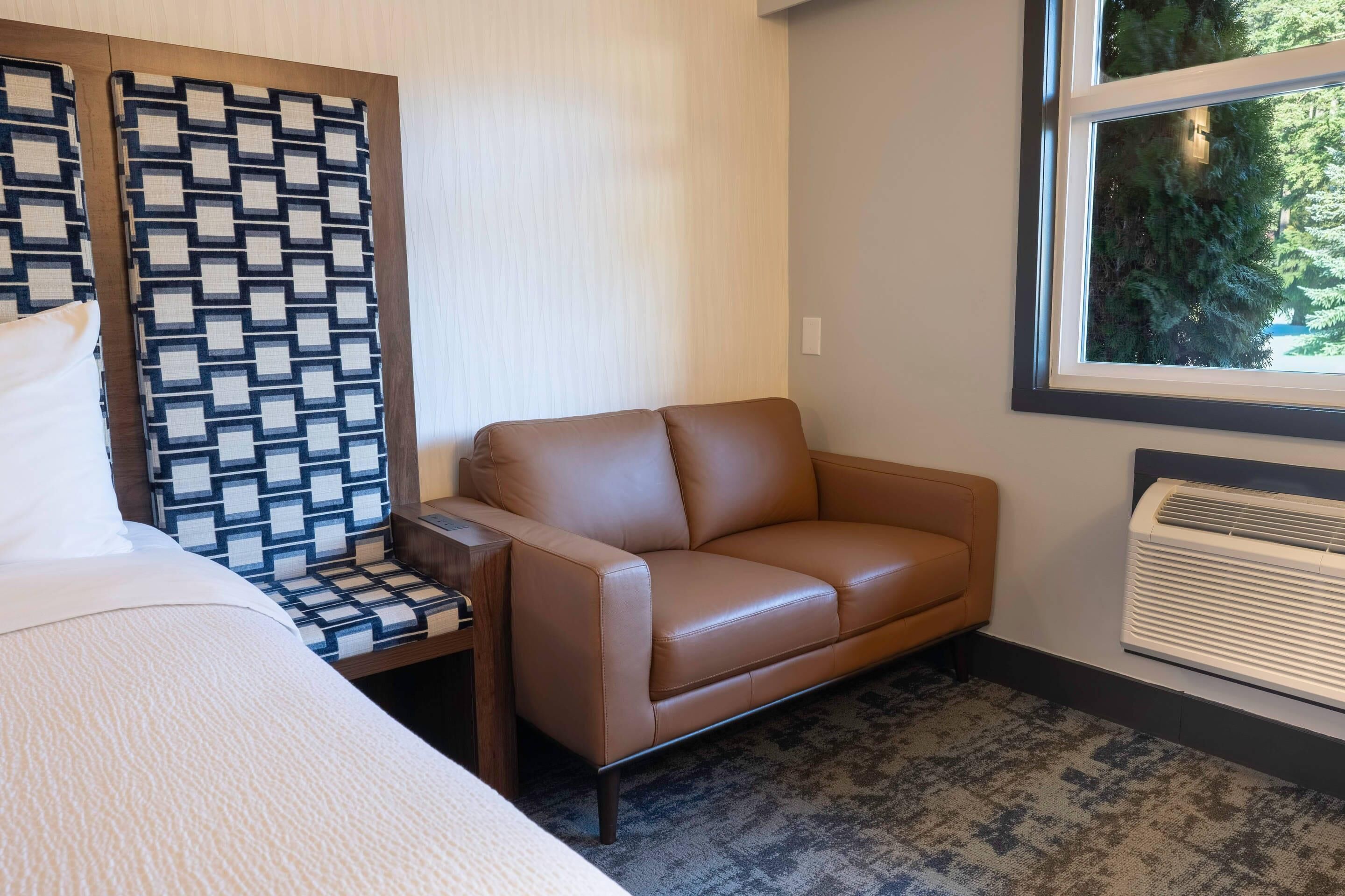 A hotel room with a brown leather sofa, a patterned chair, a bed, and a window with a view of trees.