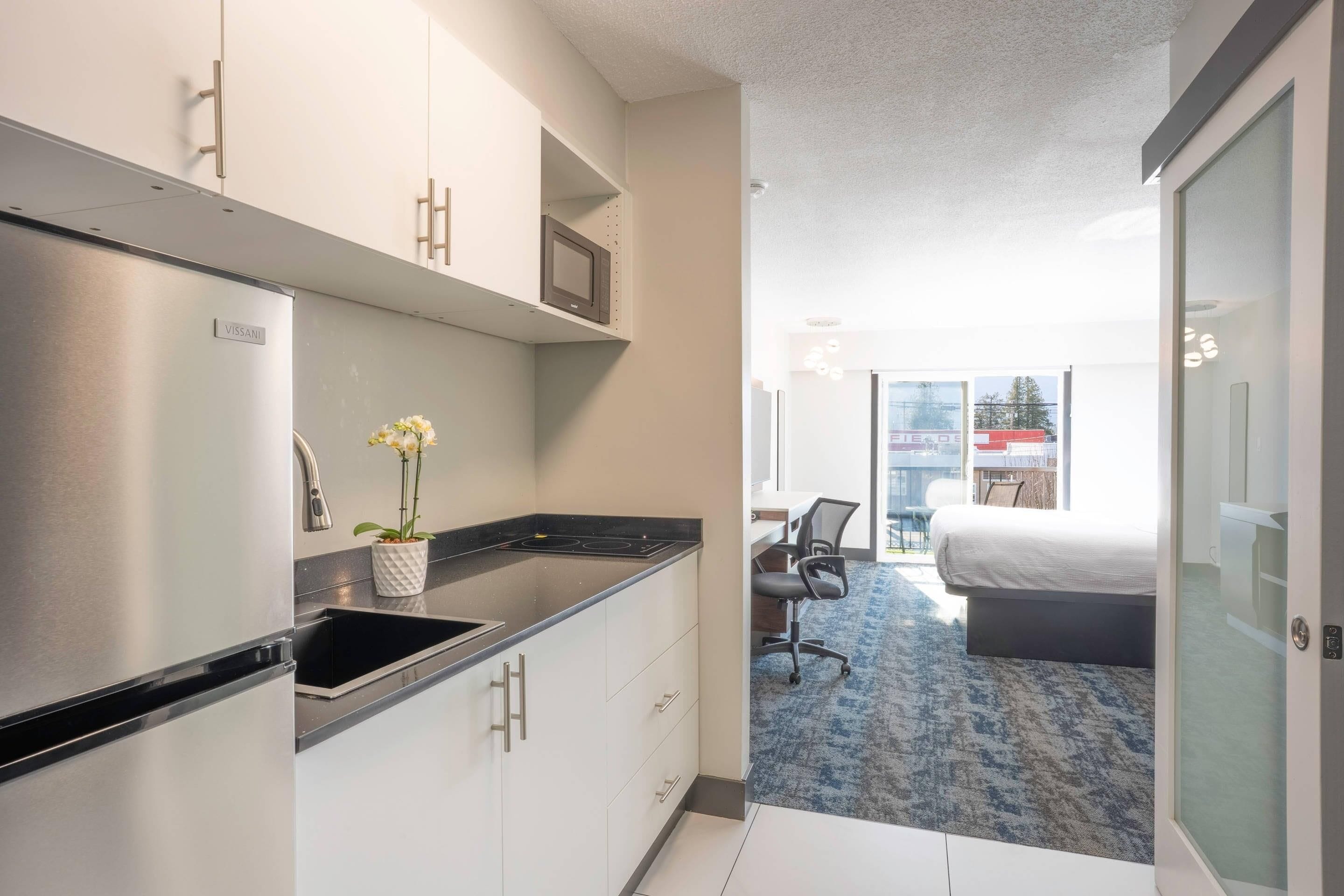 A compact hotel room with a kitchenette, a bed, a desk, and a view of the city.
