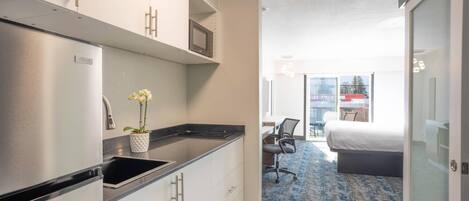 A compact hotel room with a kitchenette, a bed, a desk, and a view of the city.