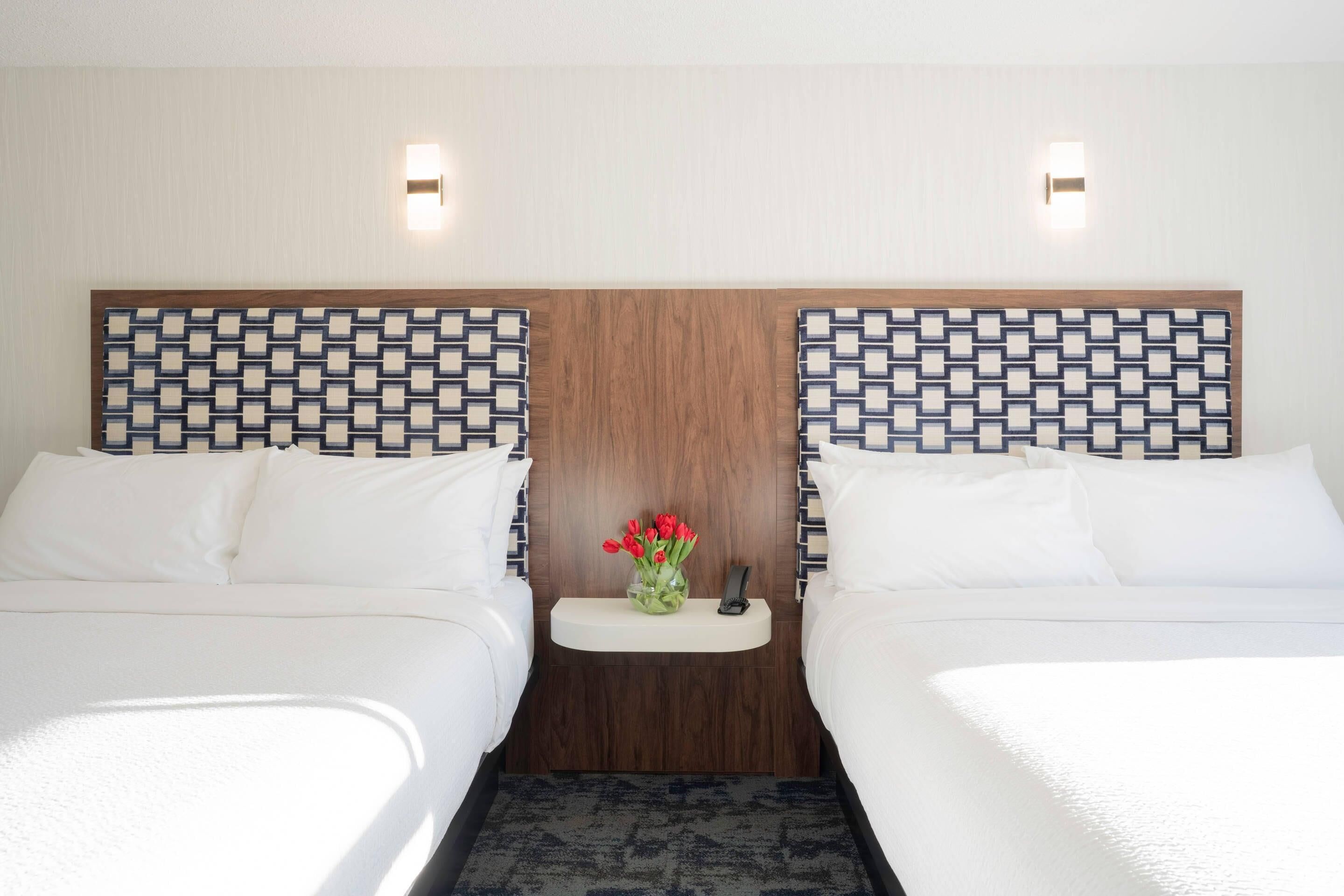 Ein Hotelzimmer mit zwei Betten, einem Holzkopfstück mit geometrischem Muster, einer kleinen Ablage mit einer Vase roter Blumen und zwei Wandleuchten.