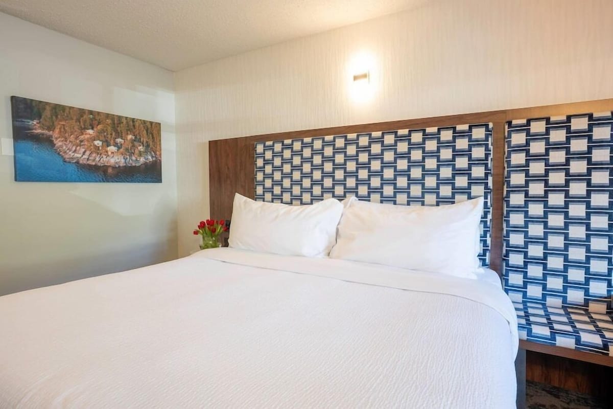 A hotel room with a large bed, a patterned headboard, a painting on the wall, and a small vase with flowers.