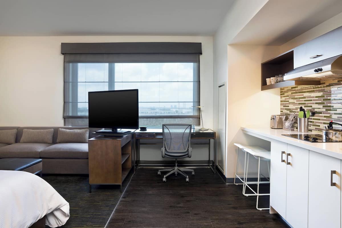 Una habitación de hotel moderna con un ventanal grande, un televisor de pantalla plana, un sofá, un escritorio con una silla y una kitchenette.