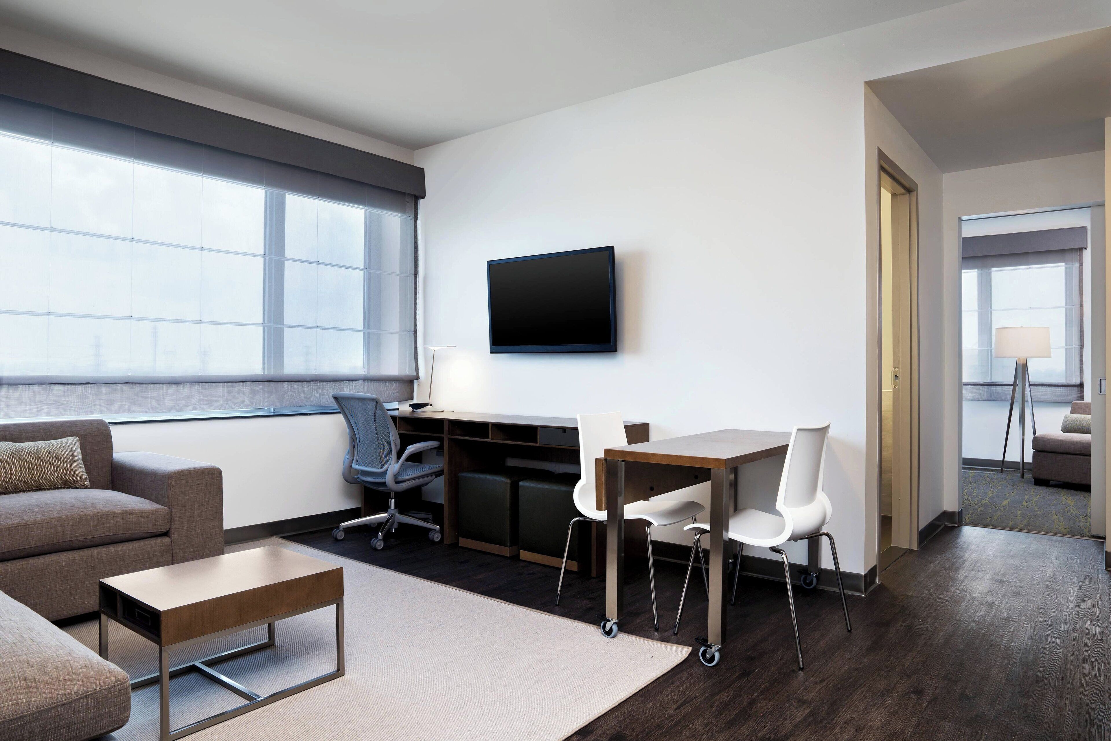 A modern hotel room with a large window, a flat-screen TV, a desk with two chairs, a sofa, and a coffee table.