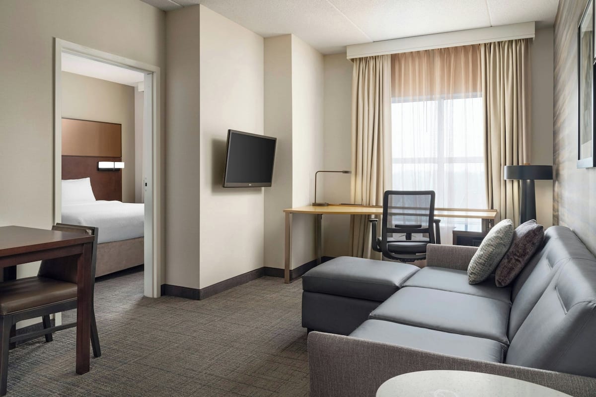 Ein Hotelzimmer mit Bett, Schreibtisch mit Stuhl, einer Sitzecke und einem Fernseher, sowie einem Fenster mit Vorhängen.