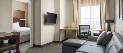 Habitación de hotel con cama, escritorio con silla, sofá, televisor y ventana con cortinas.
