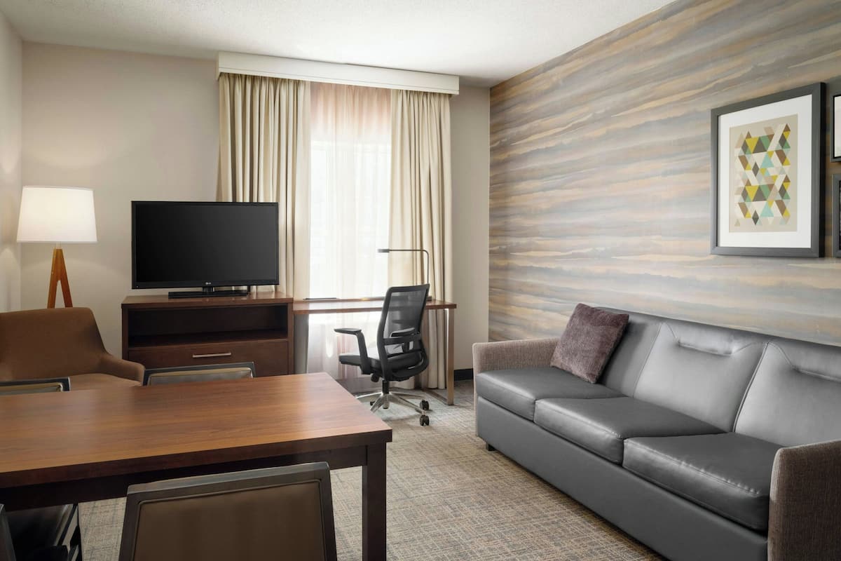 Ein Hotelzimmer mit einem Holztisch, einem Fernseher, einem grauen Sofa und einem gerahmten Bild an der Wand.