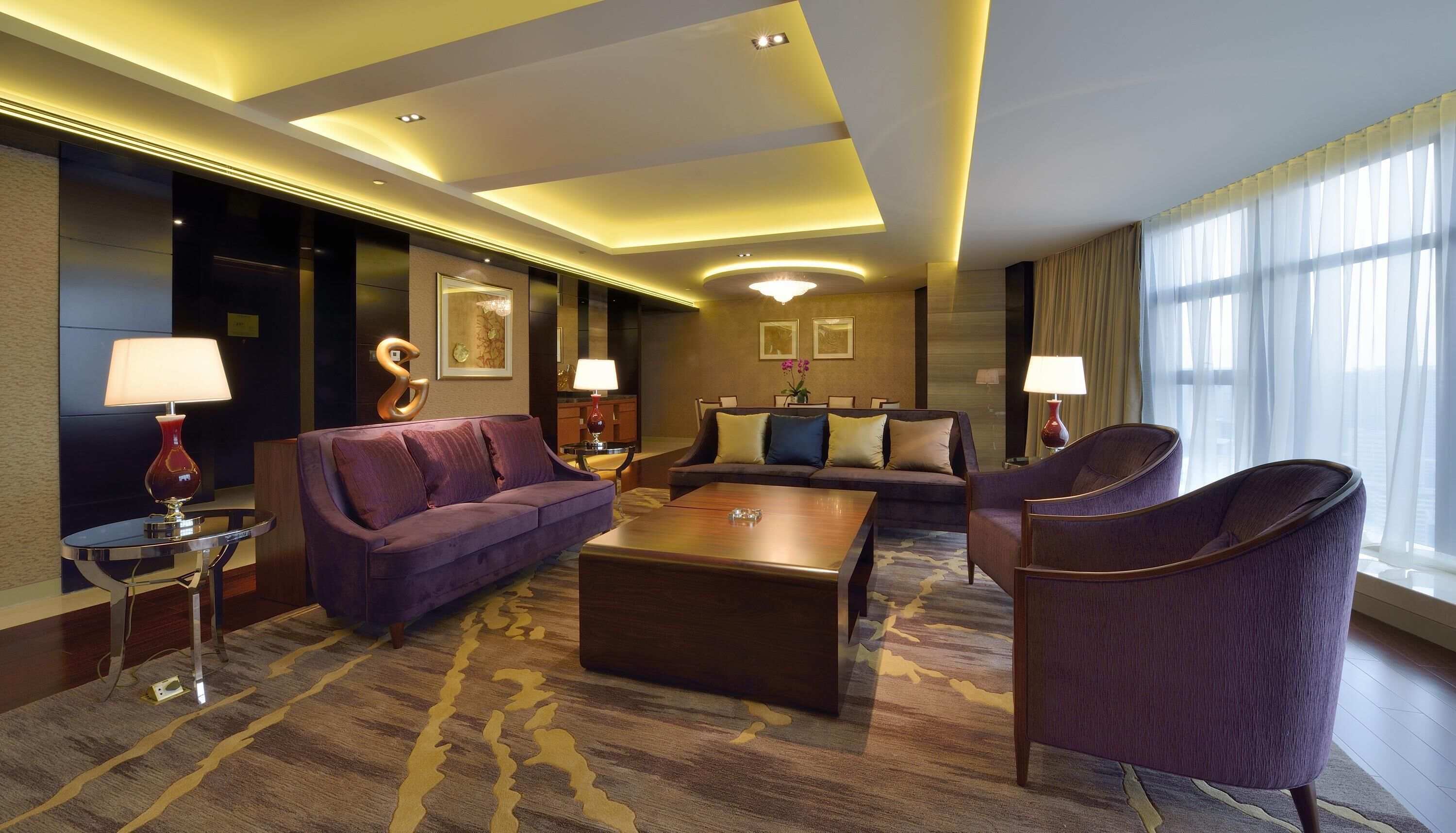 Una habitación de hotel moderna con un sofá morado, una mesa de centro de madera y un ventanal con cortinas traslúcidas.