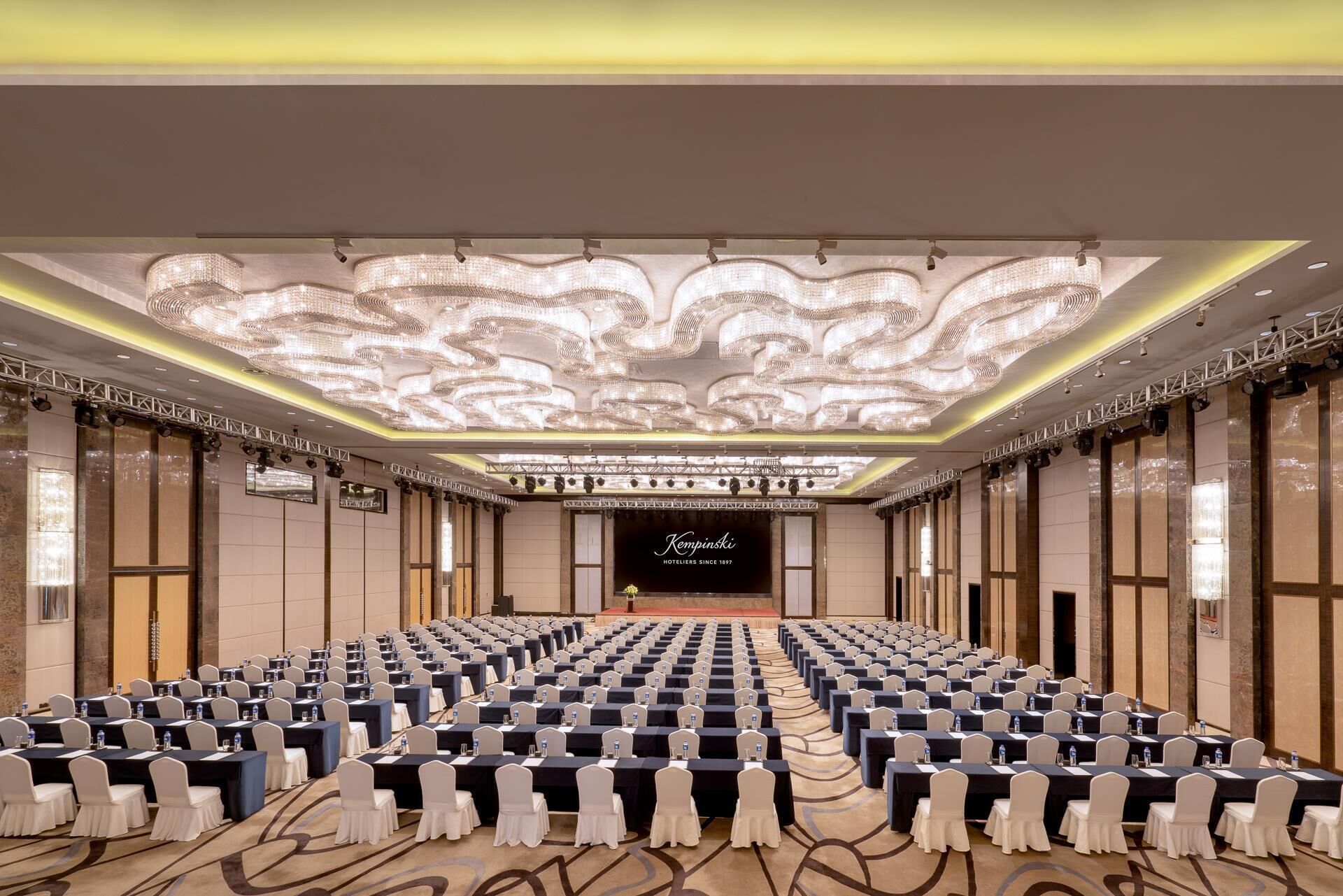 Ein großer Konferenzraum mit Tischen und Stühlen, einer Kandelaber und einem Schild mit der Aufschrift "Kempinski Hotels Since 1897".