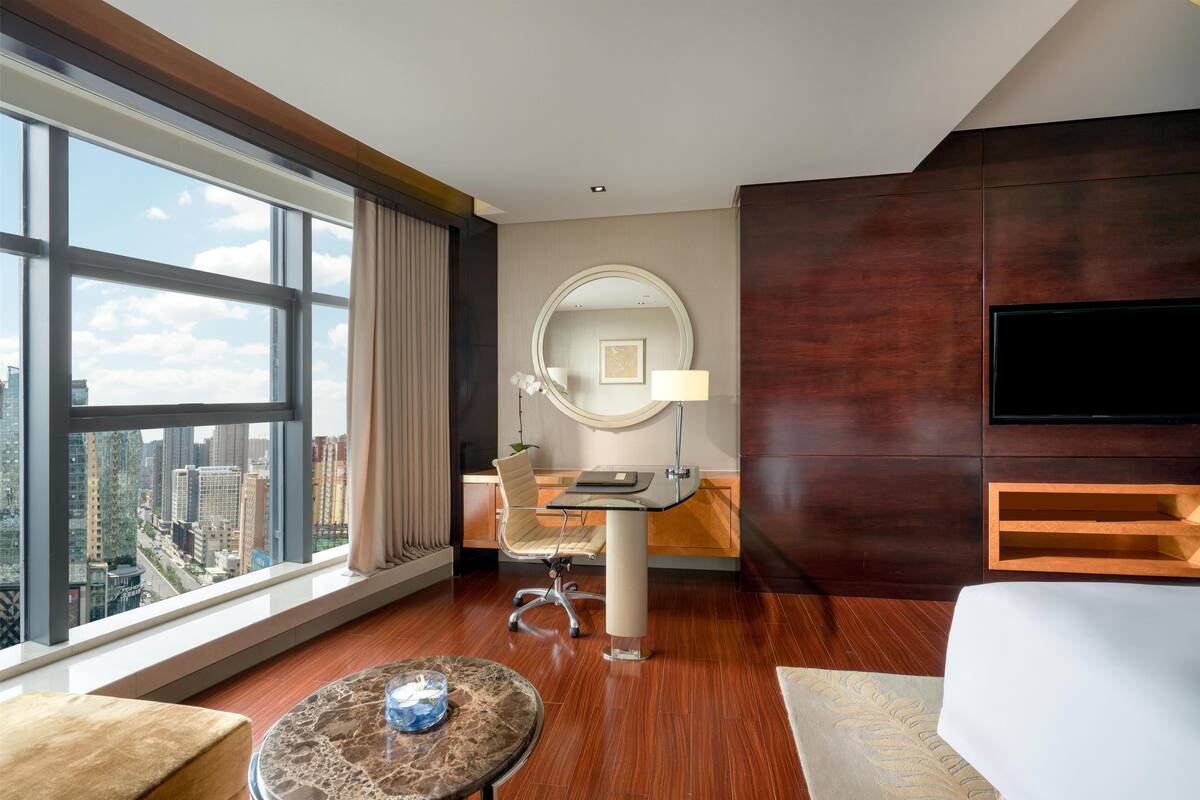 Une chambre d’hôtel avec une grande fenêtre, un miroir rond, un bureau, une chaise, une télévision et un lit.