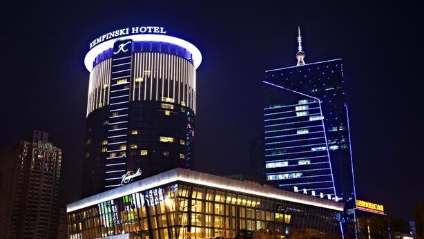 Kempinski Hotel, mit modernem Design, präsentiert einen zylindrischen Turm und ein rechteckiges Gebäude mit beleuchteten Fenstern.