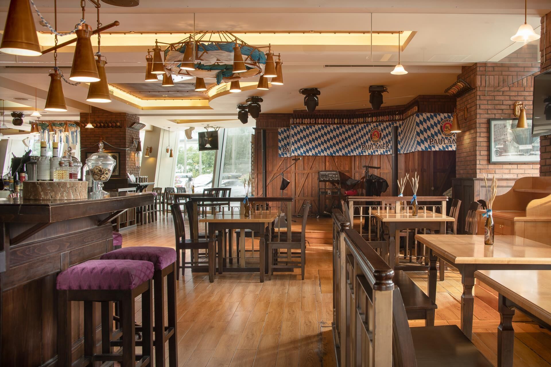A restaurant interior with wooden tables and chairs, hanging pendant lights, and a brick wall.
